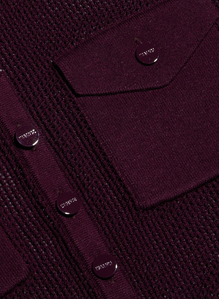 Primer plano de un top «Carine» de color burdeos oscuro con textura, que cuenta con botones metálicos brillantes y dos bolsillos funcionales con solapas, lo que resalta los detalles de su fino y ligero tejido de punto.