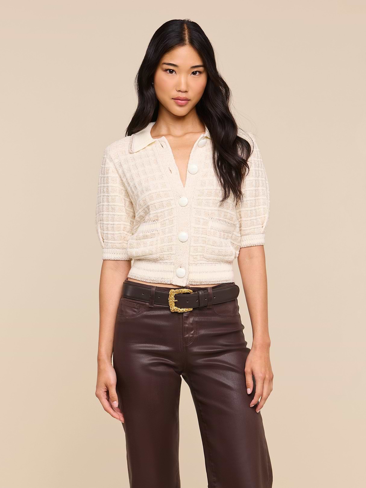A woman wears the Novalie Cardigan Top, a cream short-sleeve button-up knit with a collar and textured cardigan-inspired finish, styled with high-waisted brown leather pants and a black belt featuring a gold buckle. She stands against a plain light background.