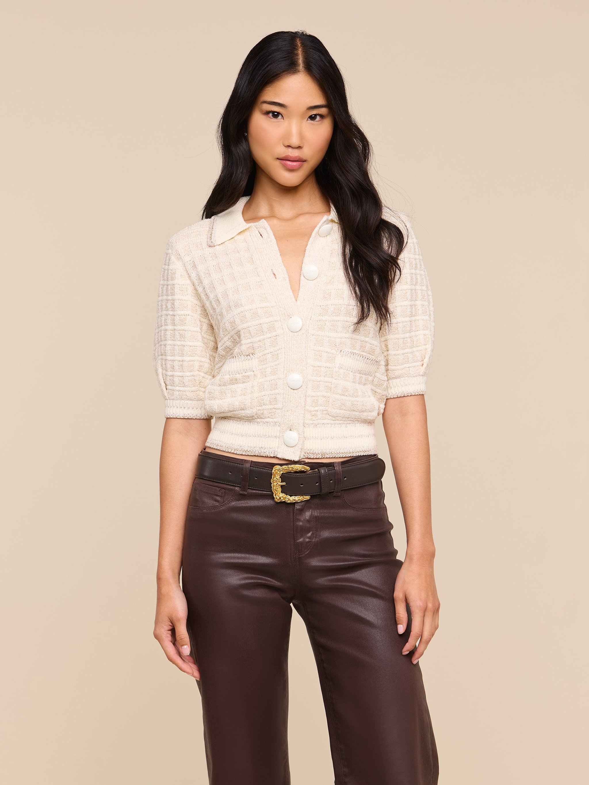 A woman wears the Novalie Cardigan Top, a cream short-sleeve button-up knit with a collar and textured cardigan-inspired finish, styled with high-waisted brown leather pants and a black belt featuring a gold buckle. She stands against a plain light background.