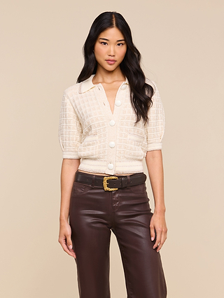 Woman with long black hair wearing the Novalie Cardigan Top—cream, textured, and buttoned at the front—paired with high-waisted brown leather pants, standing against a beige background.