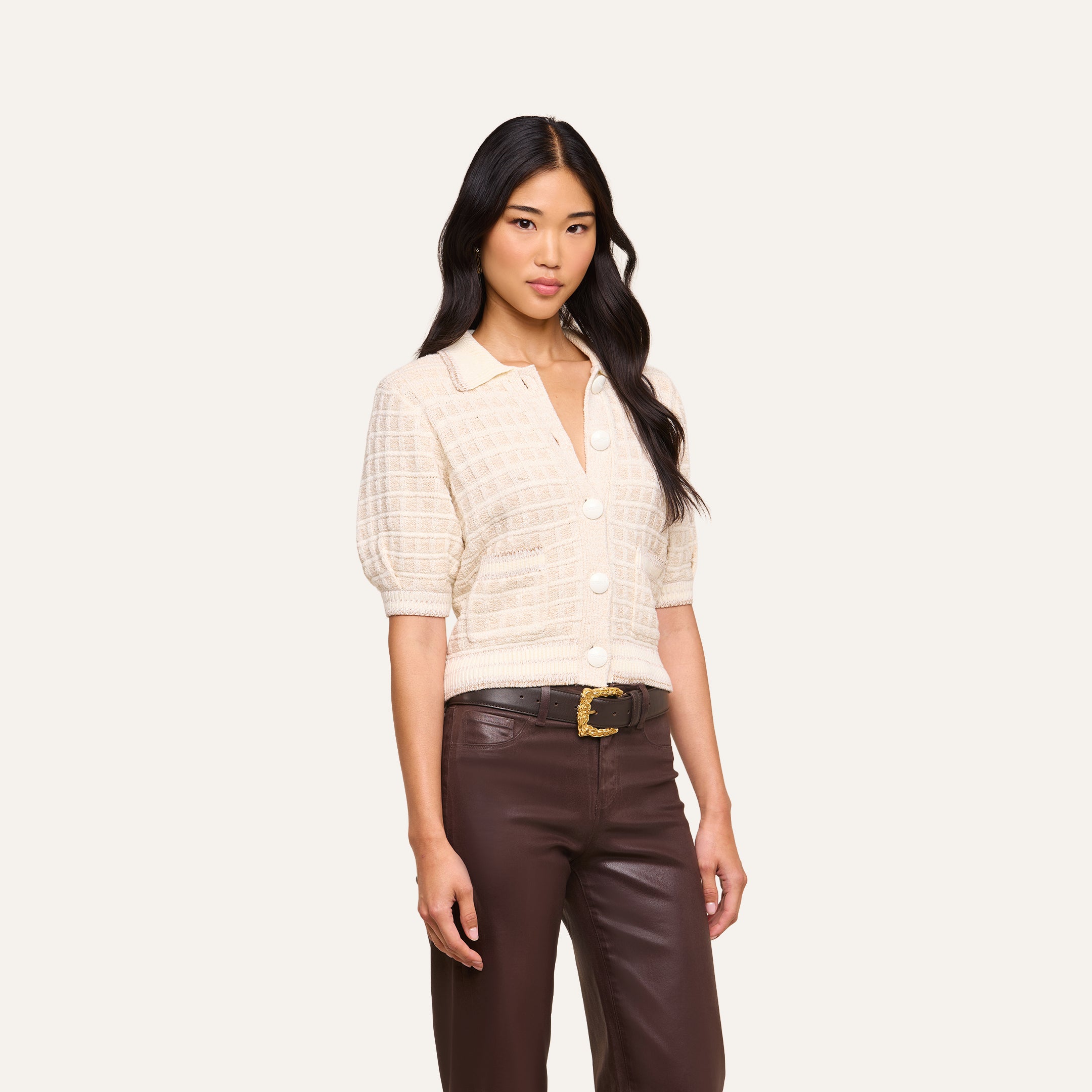 A woman with long, wavy dark hair stands against a plain light background, wearing the Novalie Cardigan Top draped over her shoulders. She pairs it with a cream short-sleeve button-up collared top, brown high-waisted pants, and a black belt with a gold buckle.