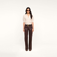 A woman with long, dark hair and a neutral expression stands against a plain white background, wearing sunglasses, the Novalie Cardigan Top—a light textured button-up shirt with elbow-length sleeves—paired with dark brown leather pants and brown shoes.