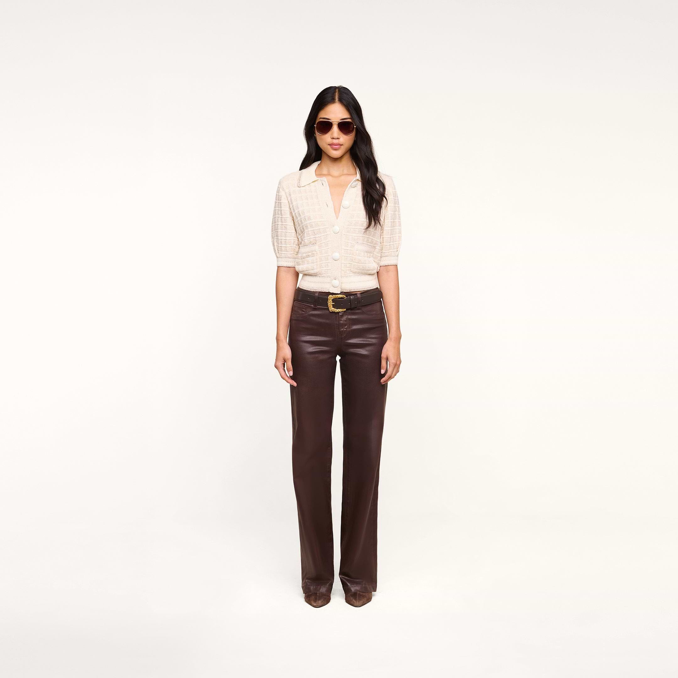 A woman with long, dark hair and a neutral expression stands against a plain white background, wearing sunglasses, the Novalie Cardigan Top—a light textured button-up shirt with elbow-length sleeves—paired with dark brown leather pants and brown shoes.