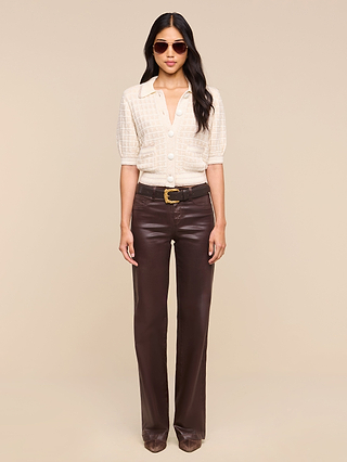 Woman standing against a beige background, wearing sunglasses, a short-sleeve cream blouse with large buttons layered under the Novalie Cardigan Top, dark brown leather pants, a black belt with a gold buckle, and brown shoes.