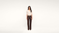 A woman wearing sunglasses, a light short-sleeve top, and dark brown pants stands facing forward against a plain white background.