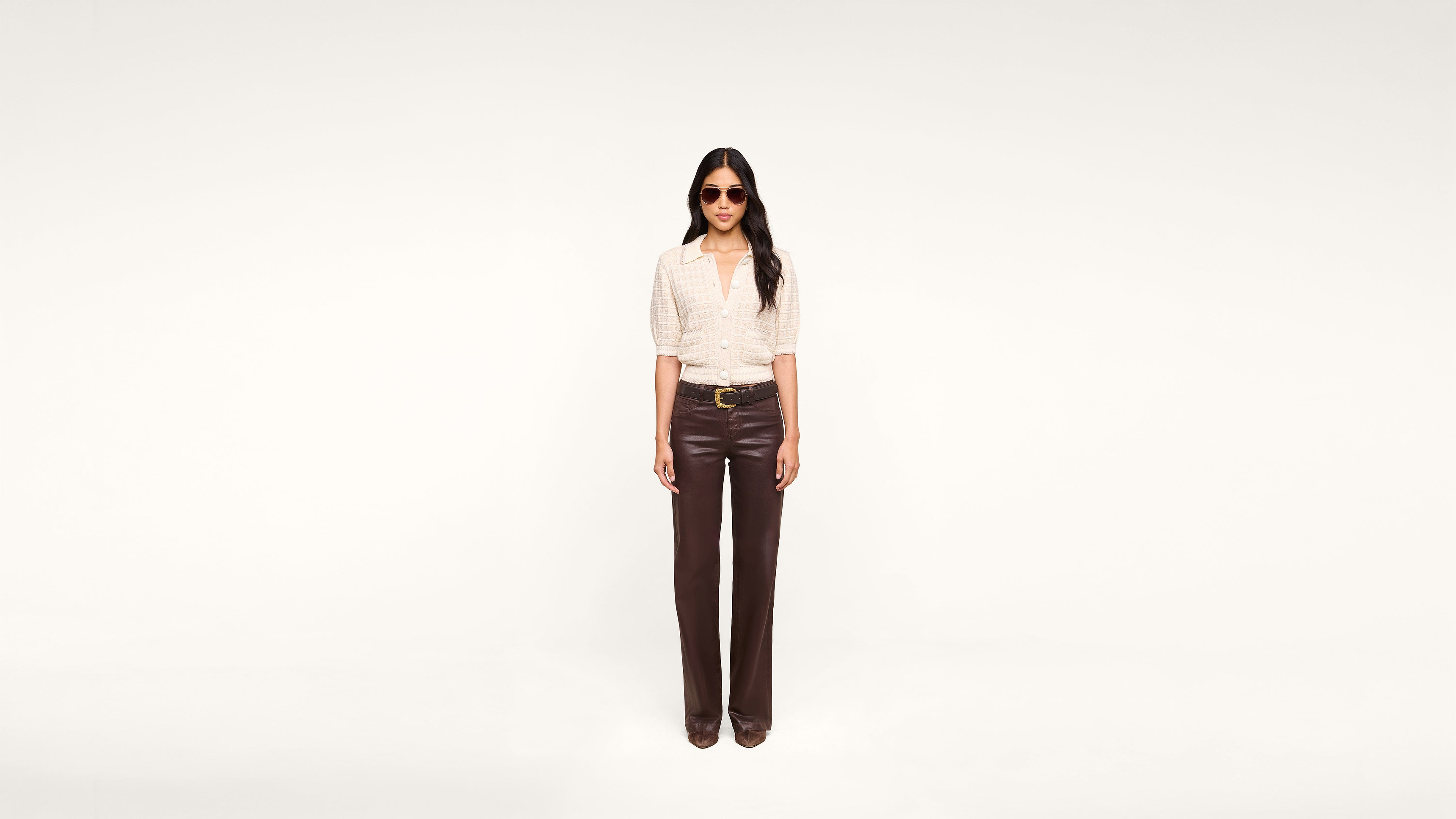 A woman wearing sunglasses, a light short-sleeve top, and dark brown pants stands facing forward against a plain white background.