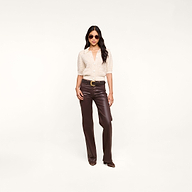 A woman wearing sunglasses, a light short-sleeve button-up shirt, and dark brown leather pants stands confidently against a plain white background, with the Novalie Cardigan Top draped elegantly over her shoulders.