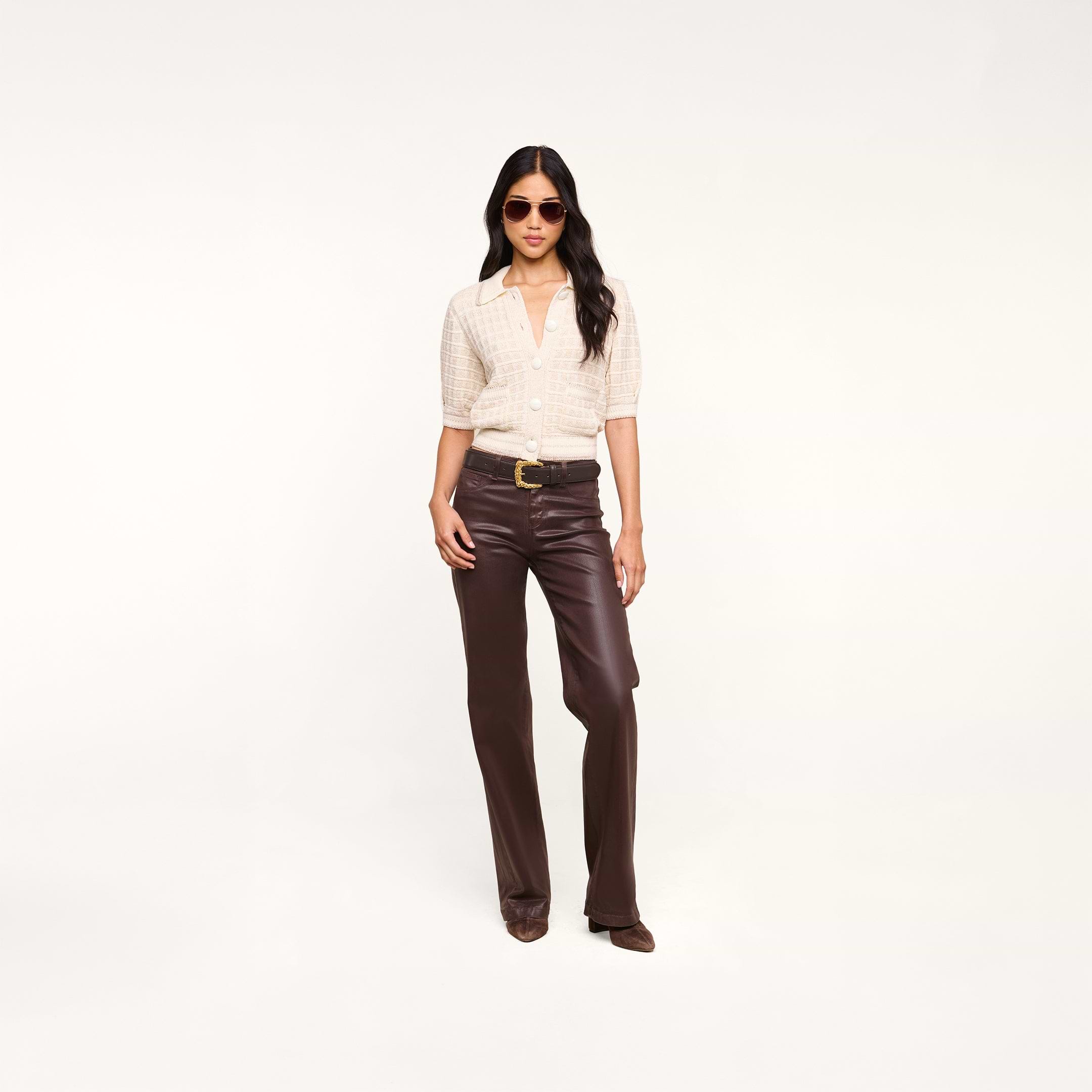 A woman wearing sunglasses, a light short-sleeve button-up shirt, and dark brown leather pants stands confidently against a plain white background, with the Novalie Cardigan Top draped elegantly over her shoulders.