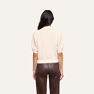 A person with long dark hair, facing away, wears the Novalie Cardigan Top—a short-sleeved cream sweater with a textured cardigan style—paired with high-waisted brown pants. The background is plain and light-colored.