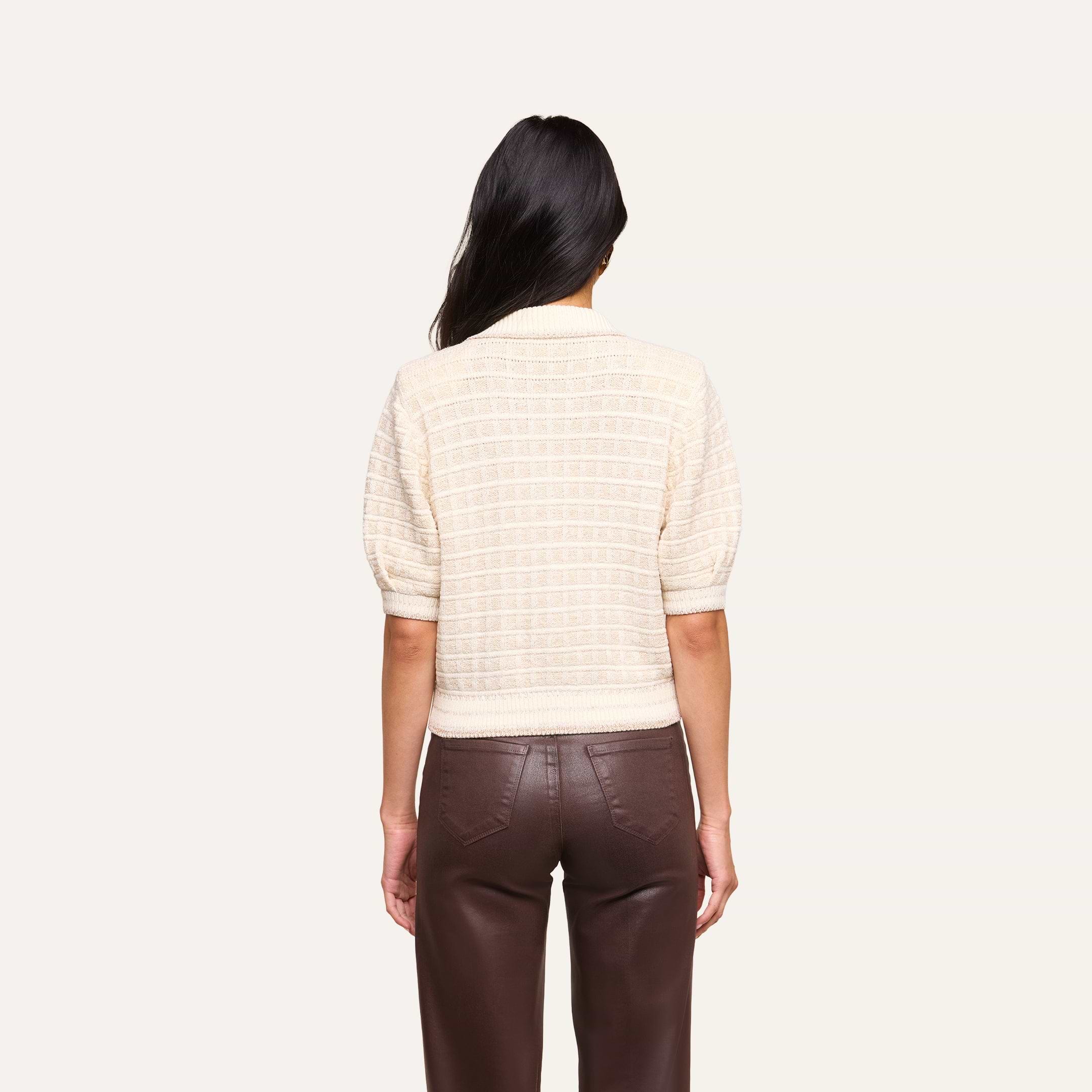 A person with long dark hair, facing away, wears the Novalie Cardigan Top—a short-sleeved cream sweater with a textured cardigan style—paired with high-waisted brown pants. The background is plain and light-colored.