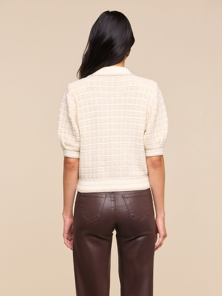 A person with long dark hair, seen from behind, wears the textured cream Novalie Cardigan Top crafted from bouclé yarn and high-waisted brown leather pants against a beige background.