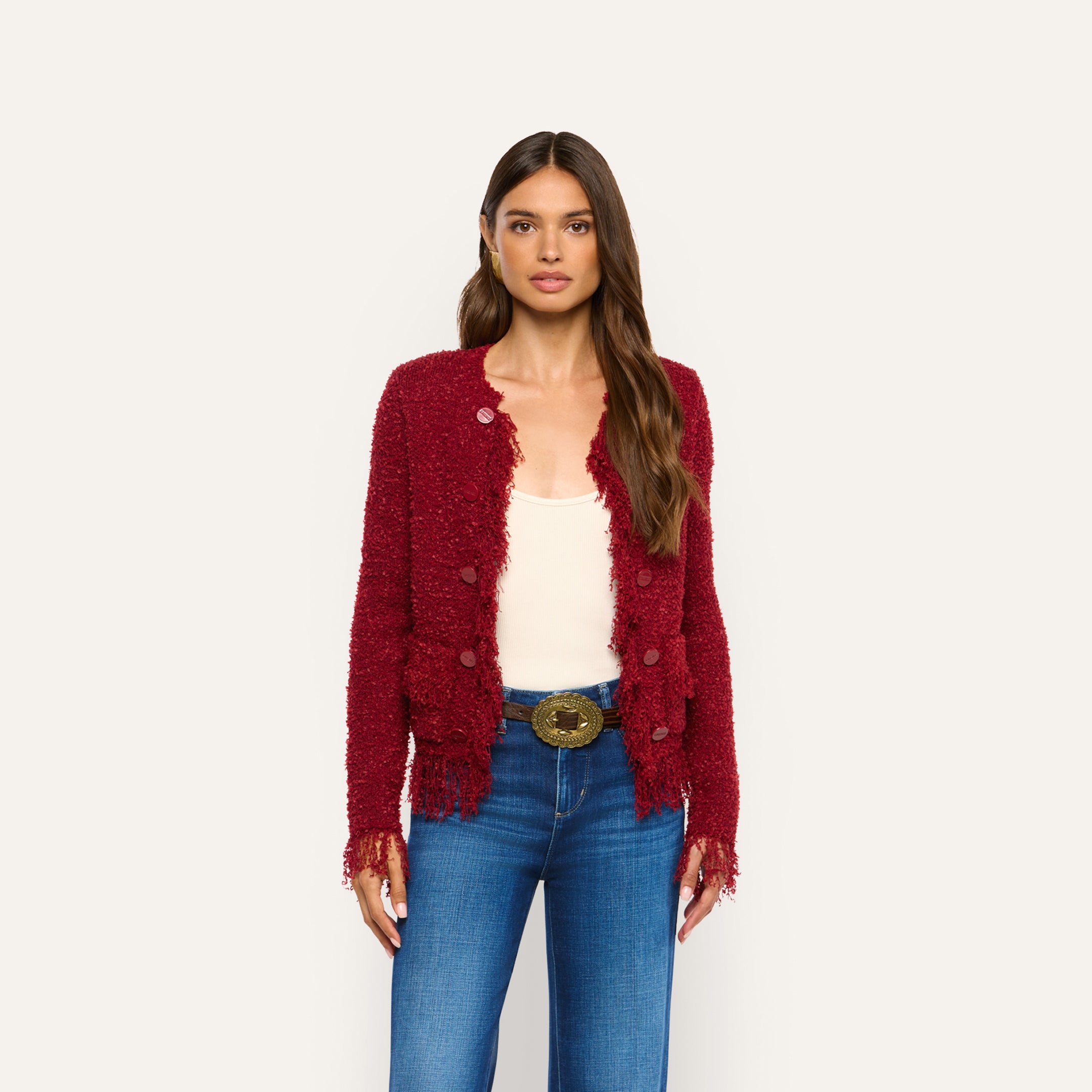 A woman wearing a cozy, textured burgundy Mireille Fringe Cardigan over a cream tank top and blue jeans with a wide decorative belt.