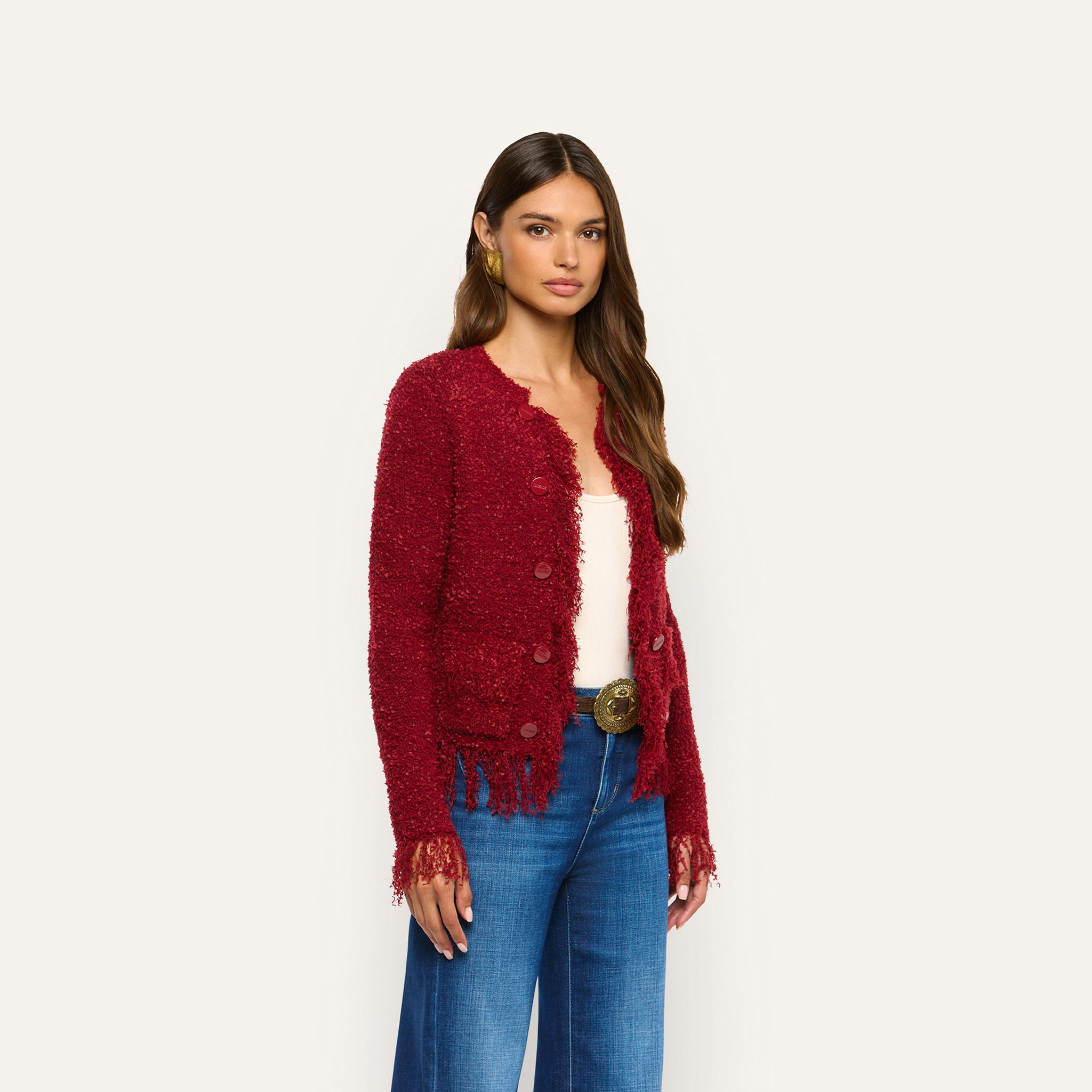 A woman wears a bold, deep red, long-sleeved Mireille Fringe Cardigan with a fringed hem and cuffs over a white top and blue jeans, creating a cozy knit look.