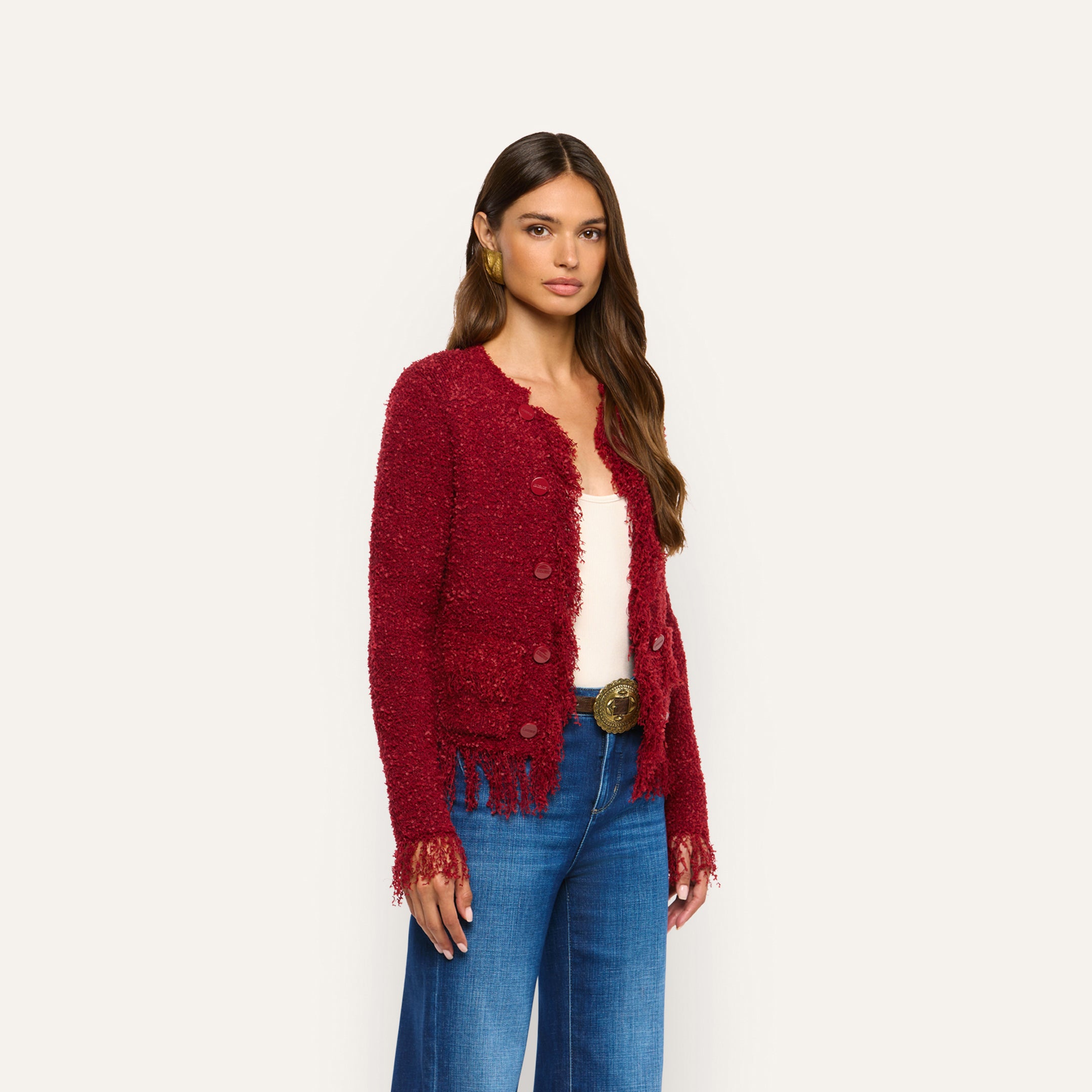 A woman wears a bold, deep red, long-sleeved Mireille Fringe Cardigan with a fringed hem and cuffs over a white top and blue jeans, creating a cozy knit look.