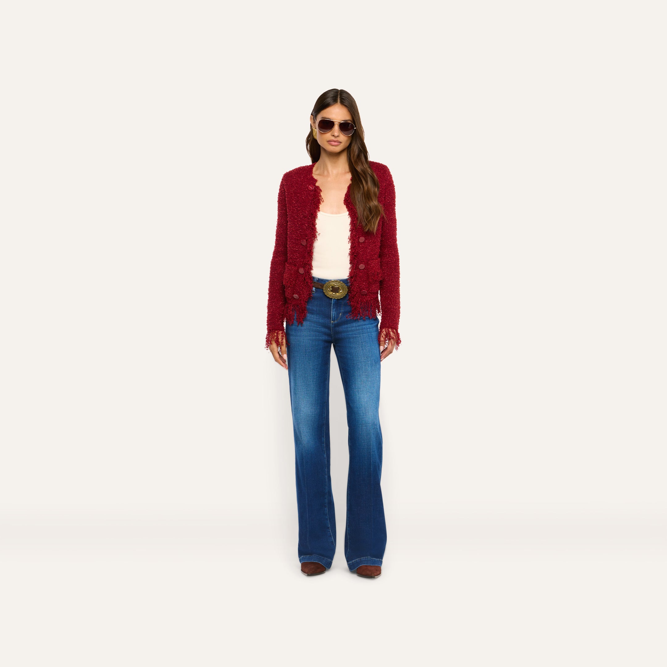 A person wears a cozy, textured burgundy Mireille Fringe Cardigan over a white top and blue flared jeans.