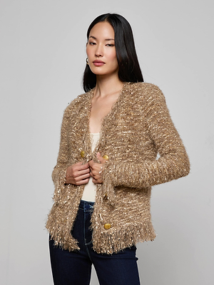 A woman with long dark hair wears the L'AGENCE Azure Fringe Cardigan Blazer, a textured gold-beige knit with fringe trim and large gold buttons, over a light top and dark jeans against a plain gray background.