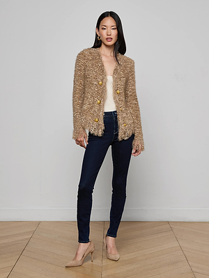 A woman with straight dark hair and a neutral expression stands against a light gray wall wearing the L'AGENCE Azure Fringe Cardigan Blazer with gold buttons, a cream top, dark blue skinny jeans, and beige heels.