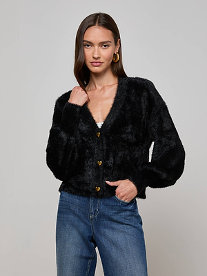 A woman wears the L'AGENCE Harriet Cardigan—cozy, black, fuzzy knit with gold heart buttons—paired with blue jeans and gold hoop earrings against a plain light gray background.