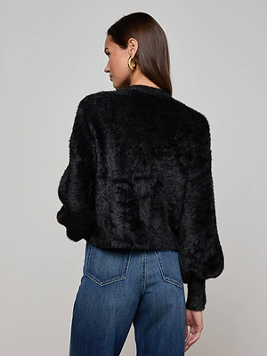 A woman with long brown hair wears the L'AGENCE Harriet Cardigan, styled with gold hoop earrings and blue jeans. She faces away from the camera, turning her head slightly against a plain light background.