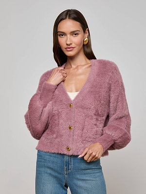 A woman with long brown hair smiles softly as she models the L'AGENCE Harriet Cardigan—a cozy-chic fuzzy knit mauve sweater with gold buttons—over a white top and blue jeans, standing against a plain gray background.