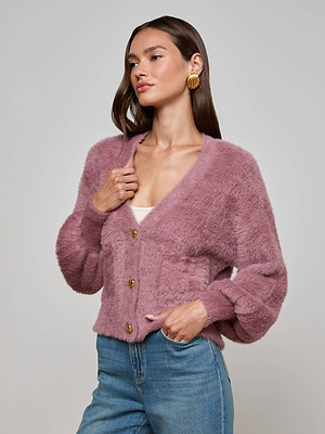 A woman with long brown hair, gold hoop earrings, and a thoughtful expression gently rests her hand on the L’AGENCE Harriet Cardigan—crafted from fuzzy knit fabric—paired with blue jeans against a plain background.