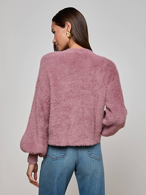 A woman with long brown hair wears gold earrings, blue jeans, and the L'AGENCE Harriet Cardigan with heart buttons, standing back to the camera against a plain light gray background.