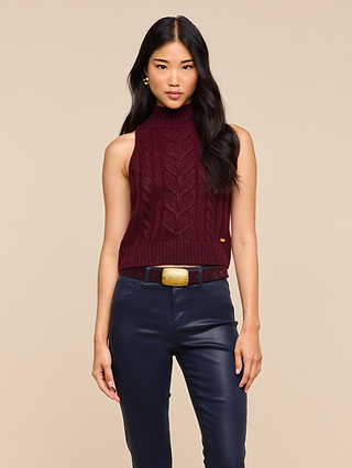 A woman with long dark hair wears the Bellini Cable-Knit Top, a wool-infused maroon turtleneck sweater, paired with navy pants and a wide belt with a gold buckle, standing against a beige background.