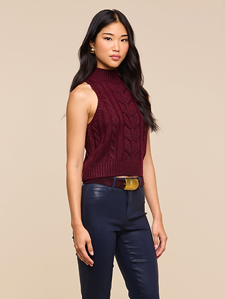 Wearing the Bellini Cable-Knit Top, a woman with long dark hair stands against a beige background, pairing the sleeveless turtleneck knit with navy pants and a wide belt.