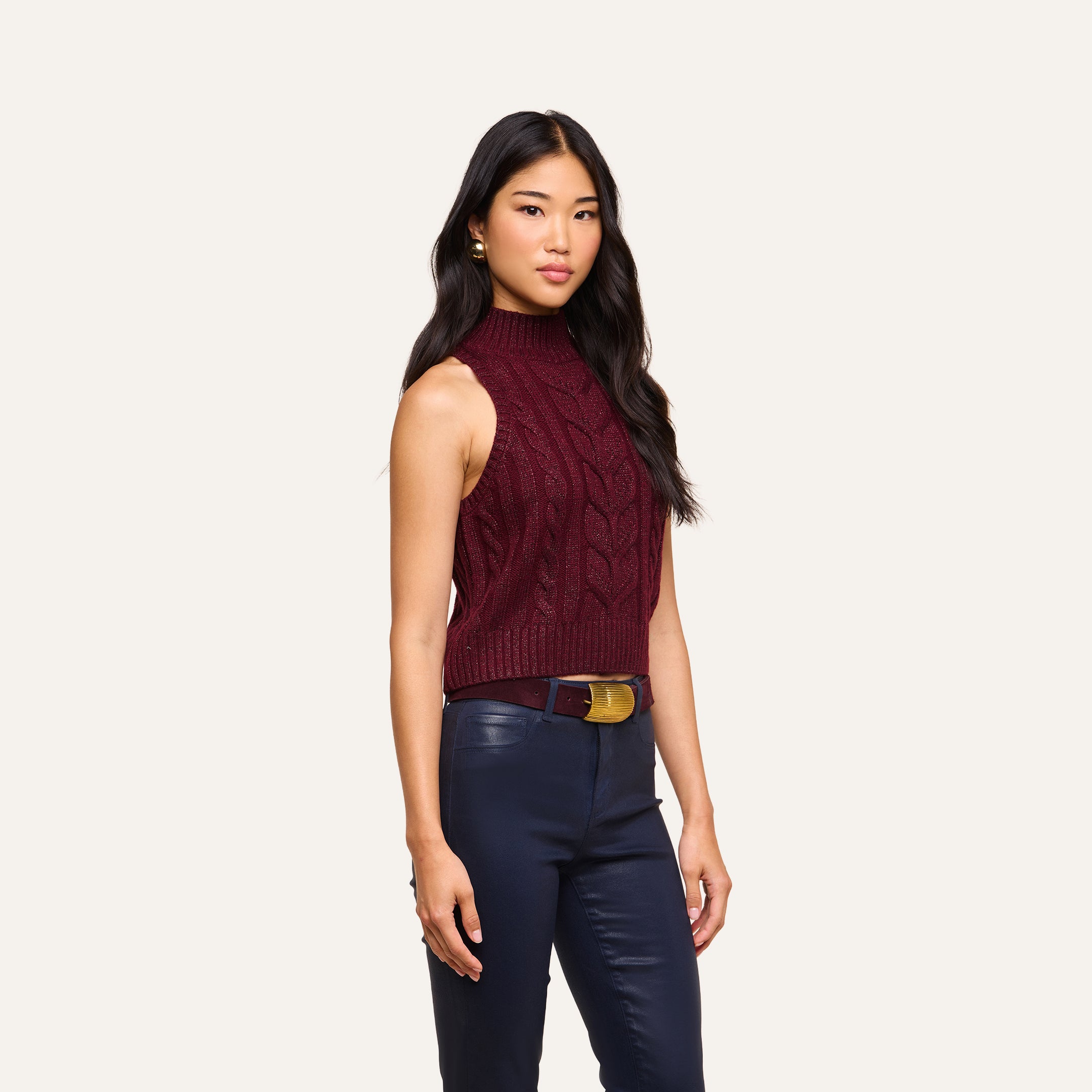 A woman stands against a plain background wearing the Bellini Cable-Knit Top and navy blue pants with a brown belt.