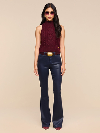 Wearing sunglasses, a woman stands against a plain beige background dressed in the Bellini Cable-Knit Top—a sleeveless maroon mock-neck tank made from wool-infused knit—paired with dark blue flared pants and burgundy shoes.