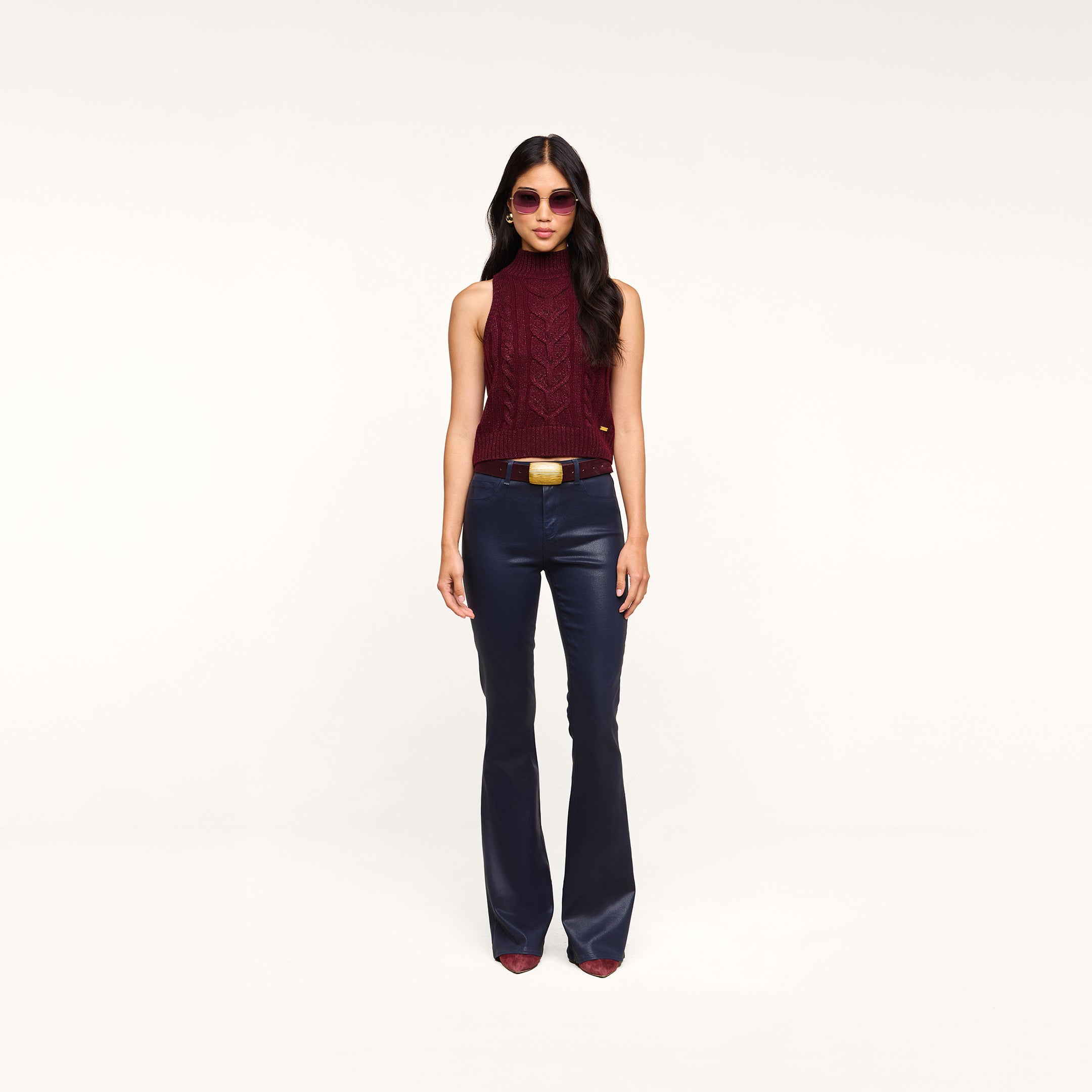 A woman stands against a plain white background, wearing sunglasses, the Bellini Cable-Knit Top in merlot red, dark blue flared pants with a belt, and maroon pointed-toe shoes.