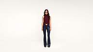 A woman wearing sunglasses, a sleeveless maroon top, dark flared pants, and heeled shoes stands against a plain white background, facing forward.