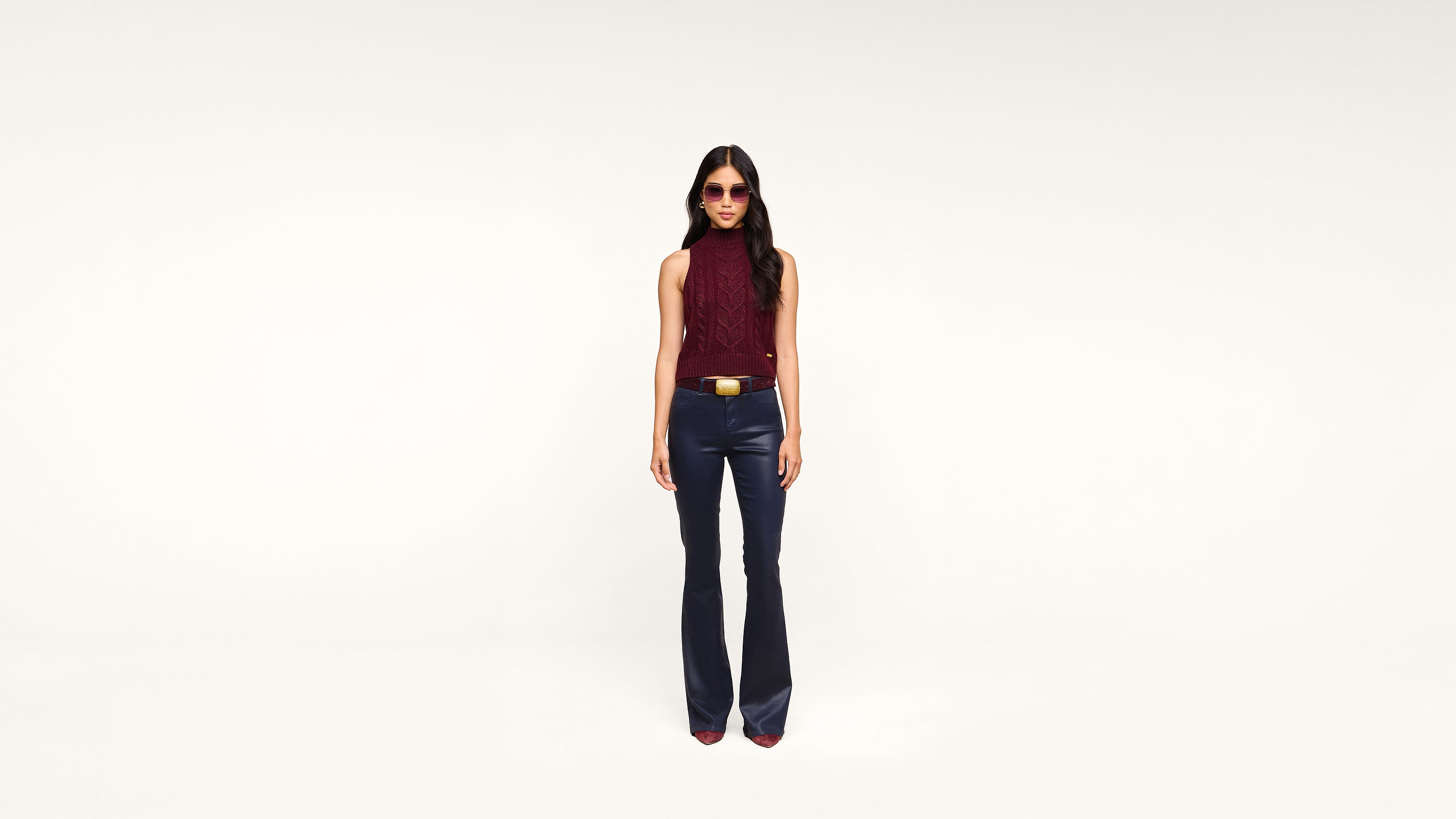 A woman wearing sunglasses, a sleeveless maroon top, dark flared pants, and heeled shoes stands against a plain white background, facing forward.