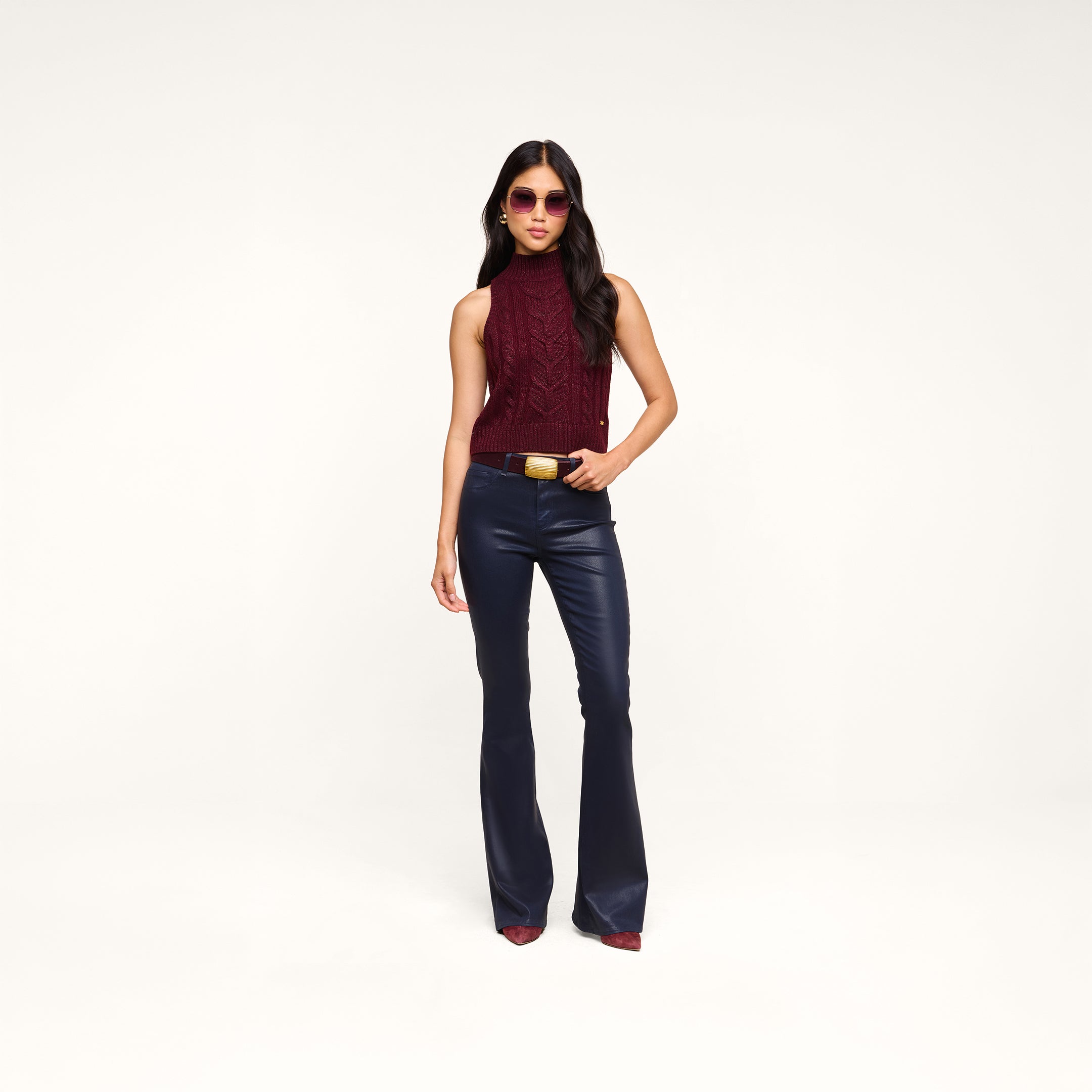 Woman wearing sunglasses, a Bellini Cable-Knit Top in merlot red, navy flared pants, and burgundy shoes stands confidently against a plain white background.