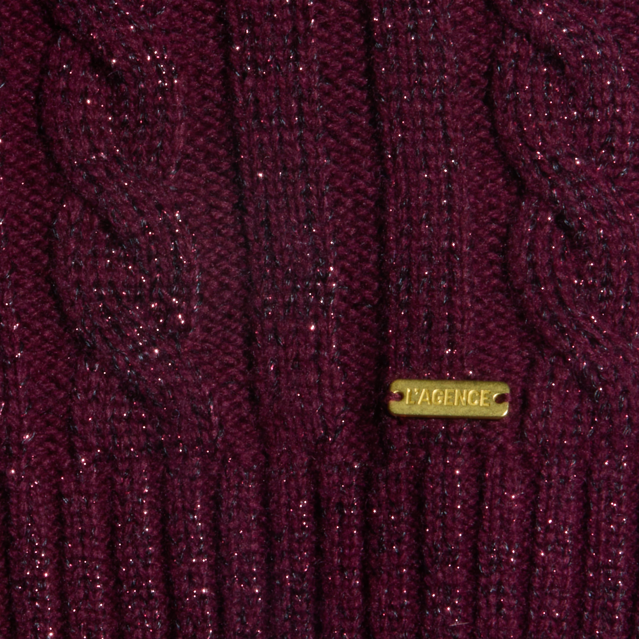 Close-up of the Bellini Cable-Knit Top in merlot red, featuring a sparkly wool-infused cable-knit fabric and a small gold rectangular tag engraved with LAGENCE.