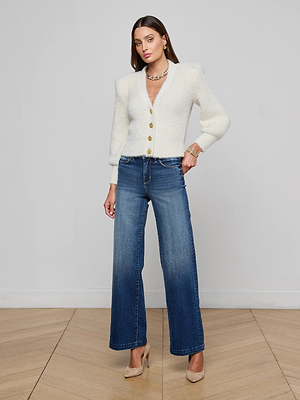 A woman poses indoors against a light wall, wearing the L’AGENCE Talulla Cardigan in white, paired with blue wide-leg jeans, beige heels, and jewelry. She stands on a wooden floor with one hand on her hip.