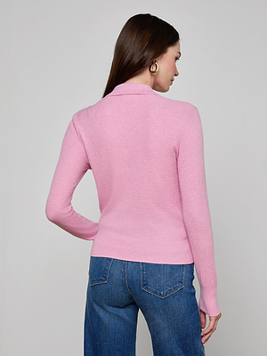 A woman with straight brown hair and gold hoop earrings wears the Sterling Sweater, a slim fit long sleeve knit pink henley top, paired with blue jeans. She is facing away against a plain light gray background.