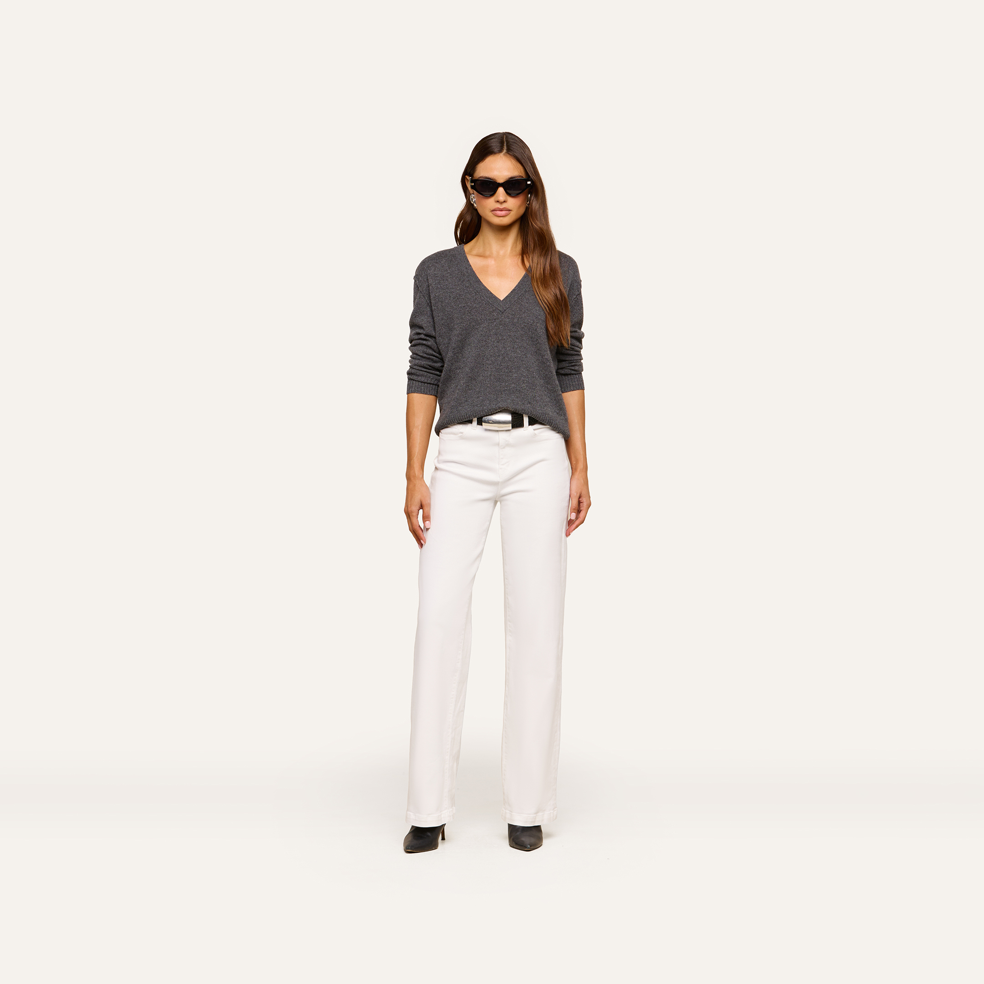 A woman wearing a gray V-neck sweater, white pants, black belt, black shoes, and sunglasses stands facing forward against a plain white background.