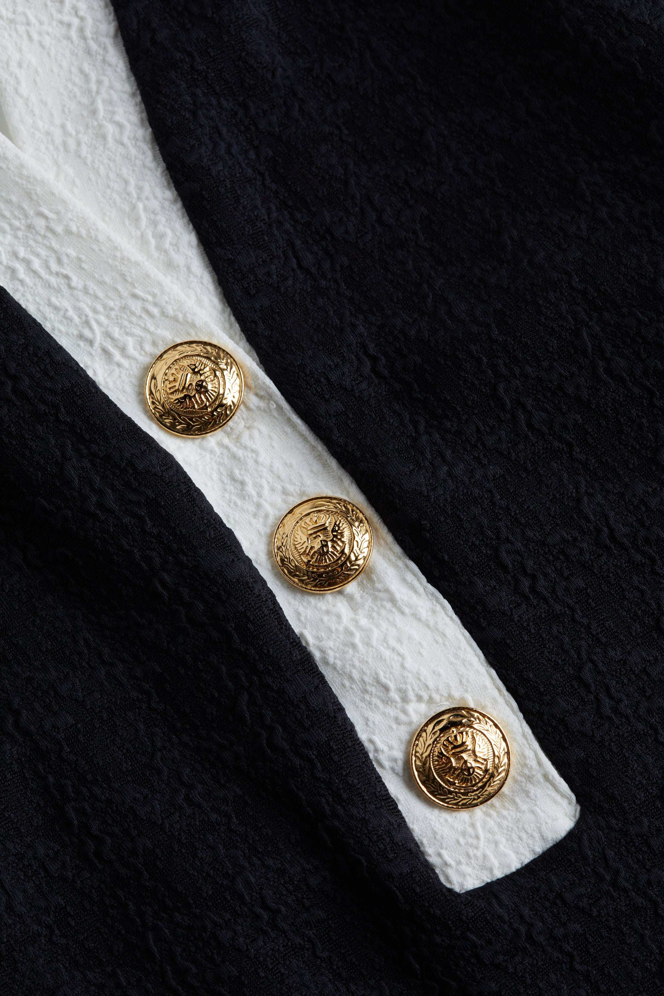 Close-up of three ornate gold buttons on the textured bouclé fabric of the Natalya Collared One-Piece, with a white stripe running underneath. The buttons feature an embossed crest design, highlighting the refined details that define this chic swimsuit.