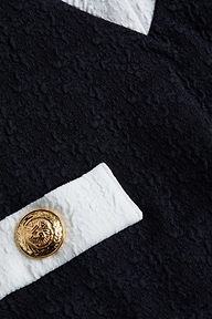A close-up of the Lisa Plunge One-Piece Swimsuit showcases textured black fabric with a white strip and a gold decorative button featuring an embossed design, evoking the look of bouclé.