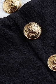 A close-up of a decorative gold button with an eagle design, featured on the Vanessa High-Waisted Bikini Bottom in textured bouclé fabric. Another matching gold button is partially visible on a white surface in the background.