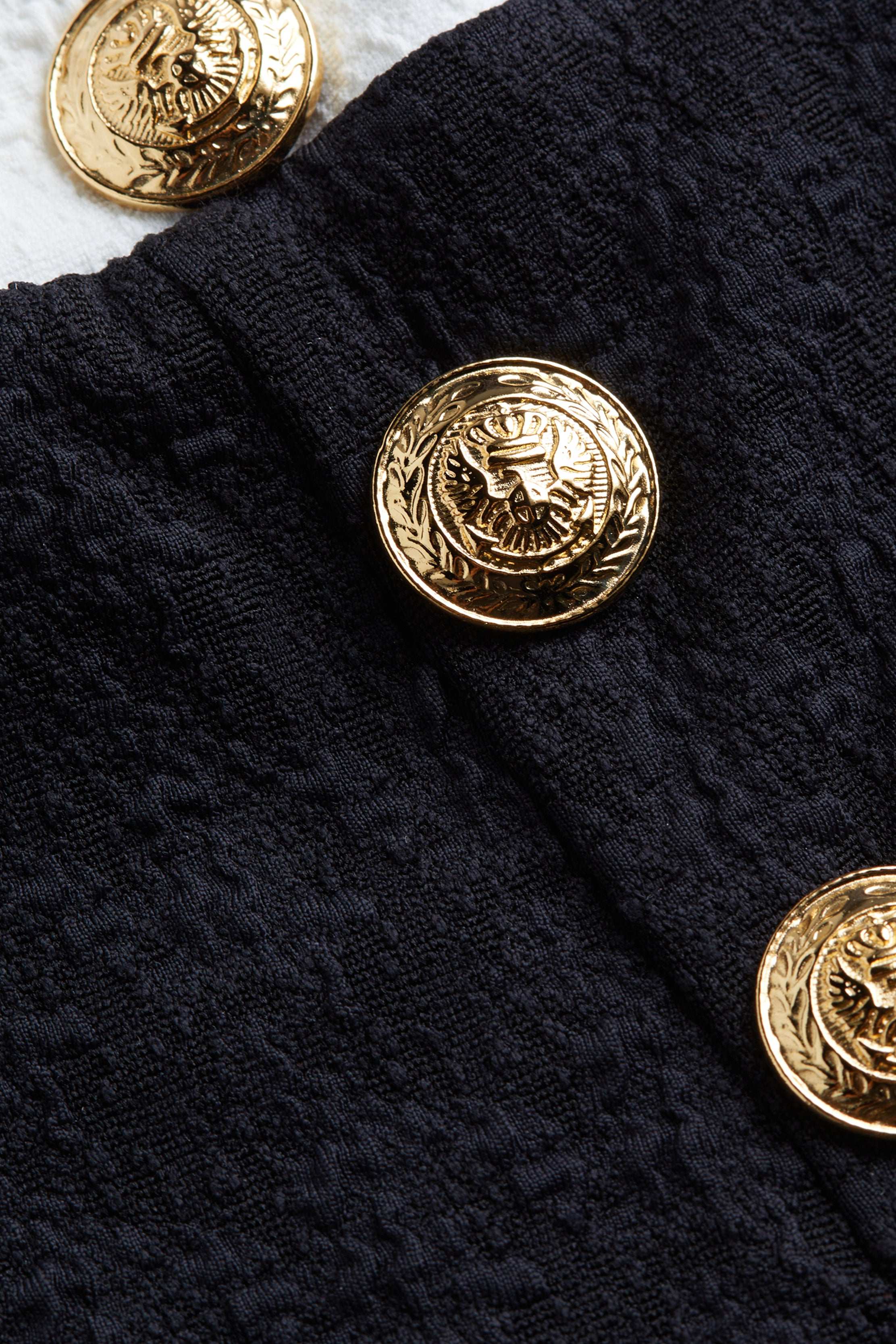 A close-up of a decorative gold button with an eagle design, featured on the Vanessa High-Waisted Bikini Bottom in textured bouclé fabric. Another matching gold button is partially visible on a white surface in the background.