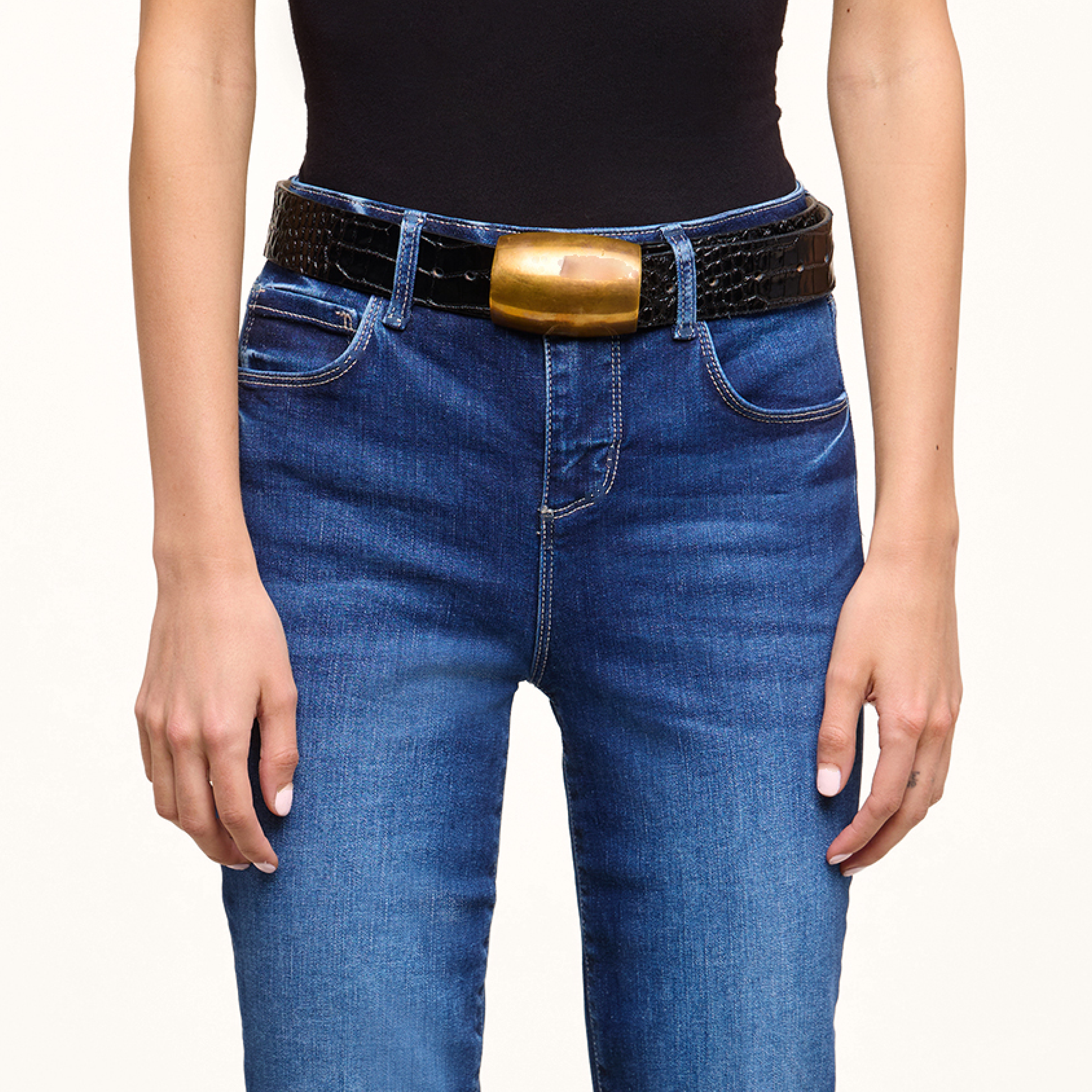 A person wearing a black top and blue jeans with a black textured belt featuring a large, rectangular gold buckle. The image shows the person from shoulders to hips, with arms relaxed at their sides.