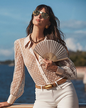 Woman in a polka dot blouse and sunglasses holding a fan by the water