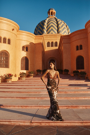 A woman in a dark, elegant dress stands on wide steps in front of a grand building with a large blue-and-gold patterned dome and warm yellow walls, bathed in golden sunlight.