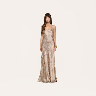 A woman with light brown wavy hair and gold bangles on her left wrist stands against a plain background, wearing the Charmaine Silk Gown—a long, sleeveless silk dress featuring adjustable straps and a metallic-patterned finish.