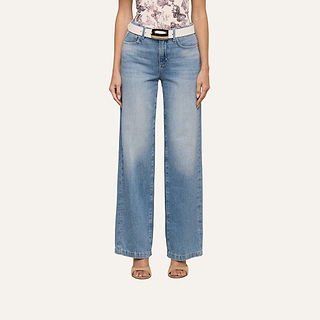 A woman stands against a plain background wearing the Scottie Wide-Leg Jean in light blue, styled with a white belt featuring a gold buckle, a floral top, and open-toe heels. Her head is not visible in the image.
