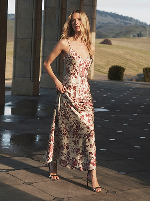 A woman in the Serita Silk Gown—a sleeveless, ankle-length white slip dress with red floral butterfly print—walks outdoors on a stone patio, green hills and columns behind her.