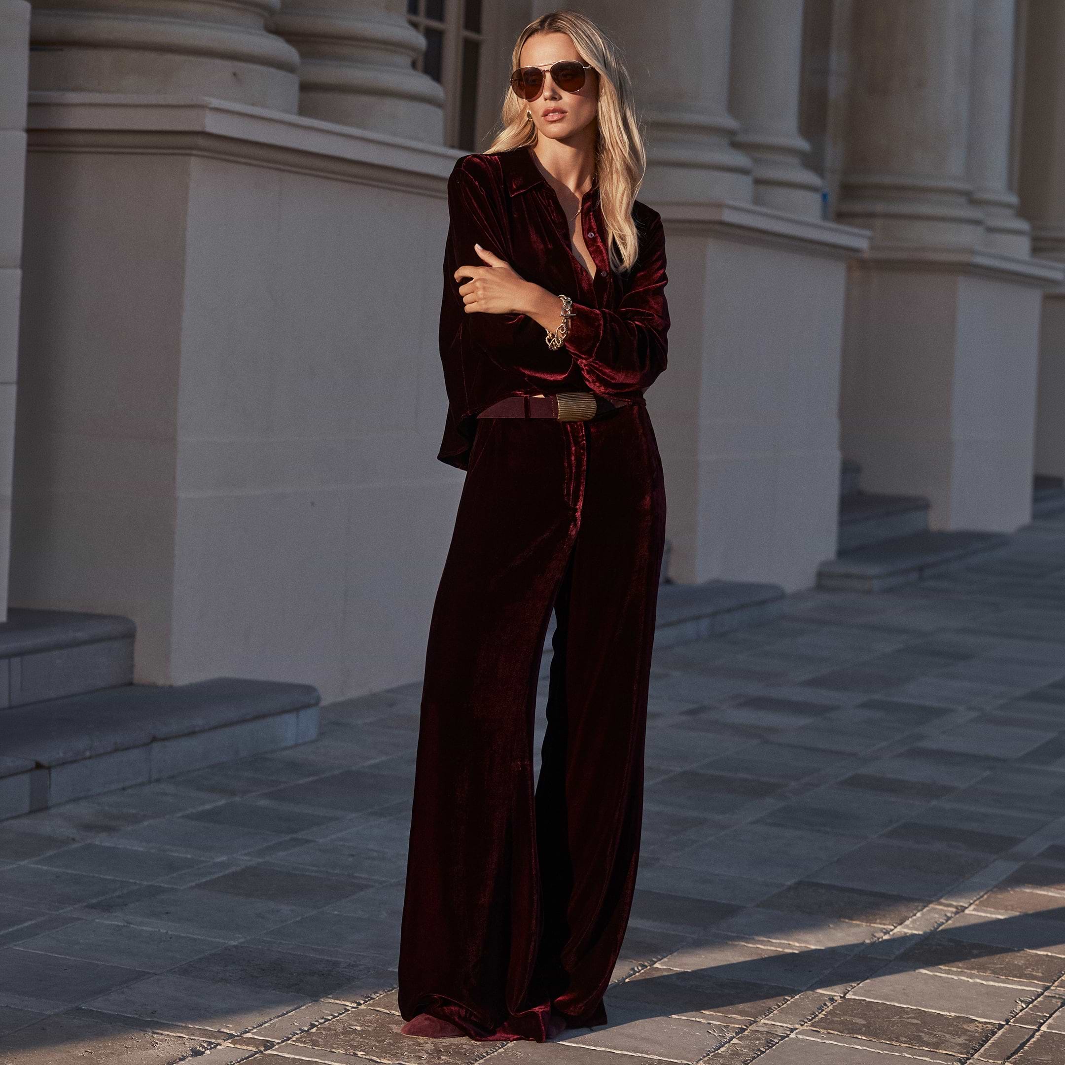 A woman wears a deep red velvet button-up shirt and matching high-rise Pilar Velvet Wide-Leg Pant.
