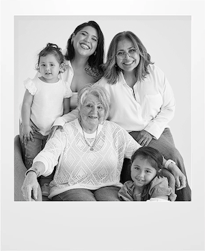 Black and white photo of five women from different generations. An elderly woman sits in front, with a young girl beside her. Behind them are two women and a young girl, all smiling warmly at the camera.